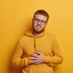 Handsome bearded man with ginger hair feels stomachache, suffers from indigestion and abdomainal cramps after eating spoiled product, wears glasses and sweatshirt, isolated on yellow background